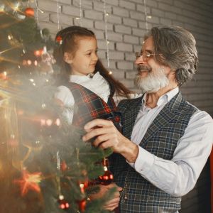 Medzigeneračná radosť pri stromčeku inšpirácia na darček v podobe jazykového kurzu Senior stojí pri vianočnom stromčeku s malou vnučkou, spoločne si užívajú sviatočnú chvíľu a radosť z darčeka v podobe jazykového kurzu.